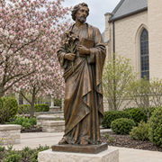 Keeper of the Lily bronze Saint Joseph sculpture holding a lily and book in an Indiana parish garden, low limestone pedestal, flowering crabapple and cream brick chapel, spring overcast.