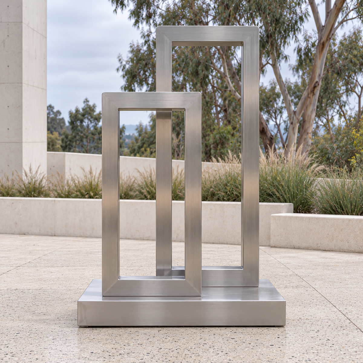 Measured Portals Brushed Stainless Steel Offset Rectangular Frames sculpture, 180 degree view in Santa Cruz museum sculpture terrace with cream concrete, ornamental grasses and eucalyptus beyond.